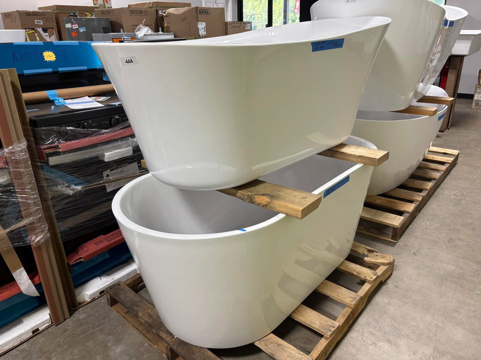 Two white freestanding acrylic soaking tubs stacked on wooden pallets inside the warehouse