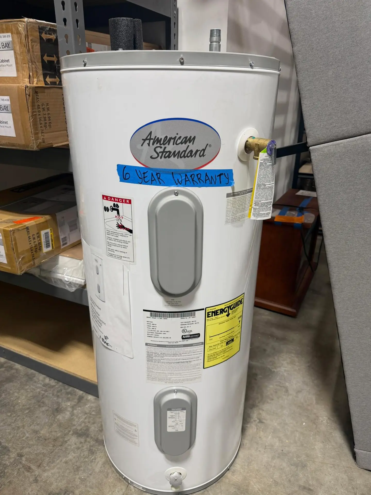 New white American Standard electric water heater with a handwritten six-year warranty label on the tank, on the warehouse floor